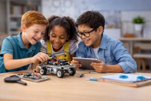 Three children smiling and working together on a robotics project at a table. One boy adjusts parts on a wheeled robot, another girl observes, and a third boy holds a tablet. They appear engaged and happy in a classroom setting.
